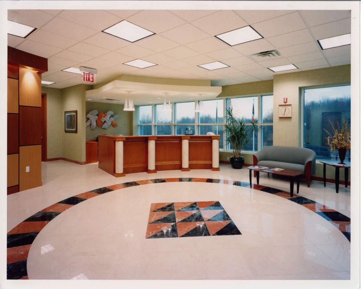 Elegant office reception area with a wooden front desk, marble flooring, and modern seating, illuminated by large windows.