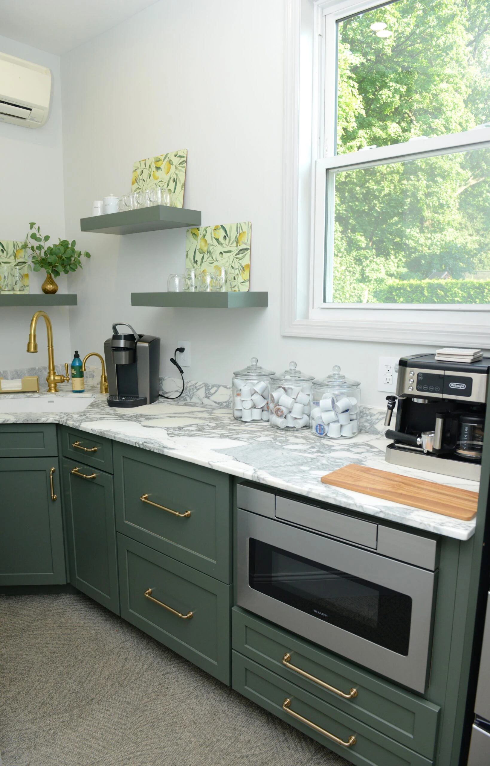 Office staff kitchen with green Shaker-style cabinets, marble countertops, brass hardware, coffee station setup, built-in microwave, floating shelves, and a functional office space design.