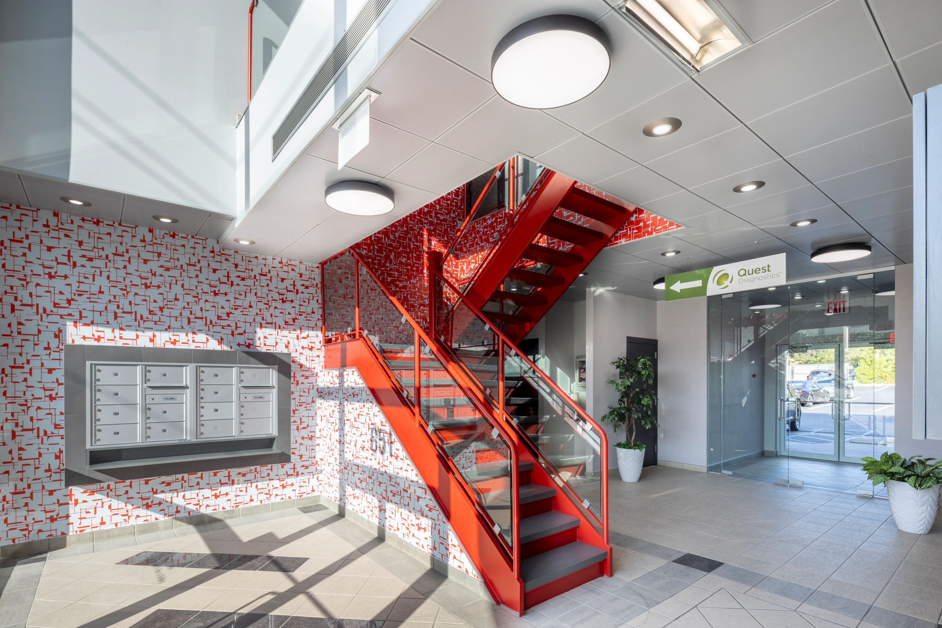 Staten Island lobby with a bold red staircase, artistic wall patterns, glass elements, geometric ceiling, modern lighting, mailbox unit, and greenery, combining art and function.