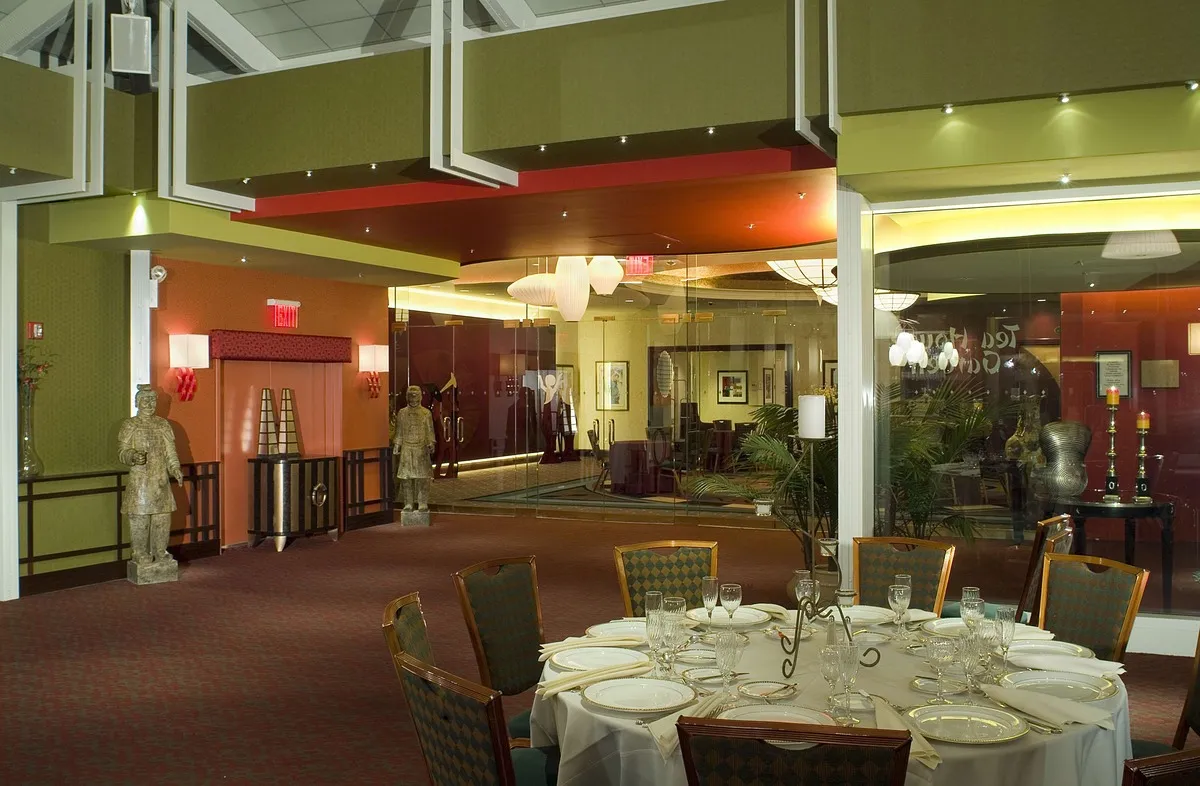 Elegant banquet seating area with a round dining table, fine china, and statues of ancient warriors, leading into a modern glass-enclosed lounge.