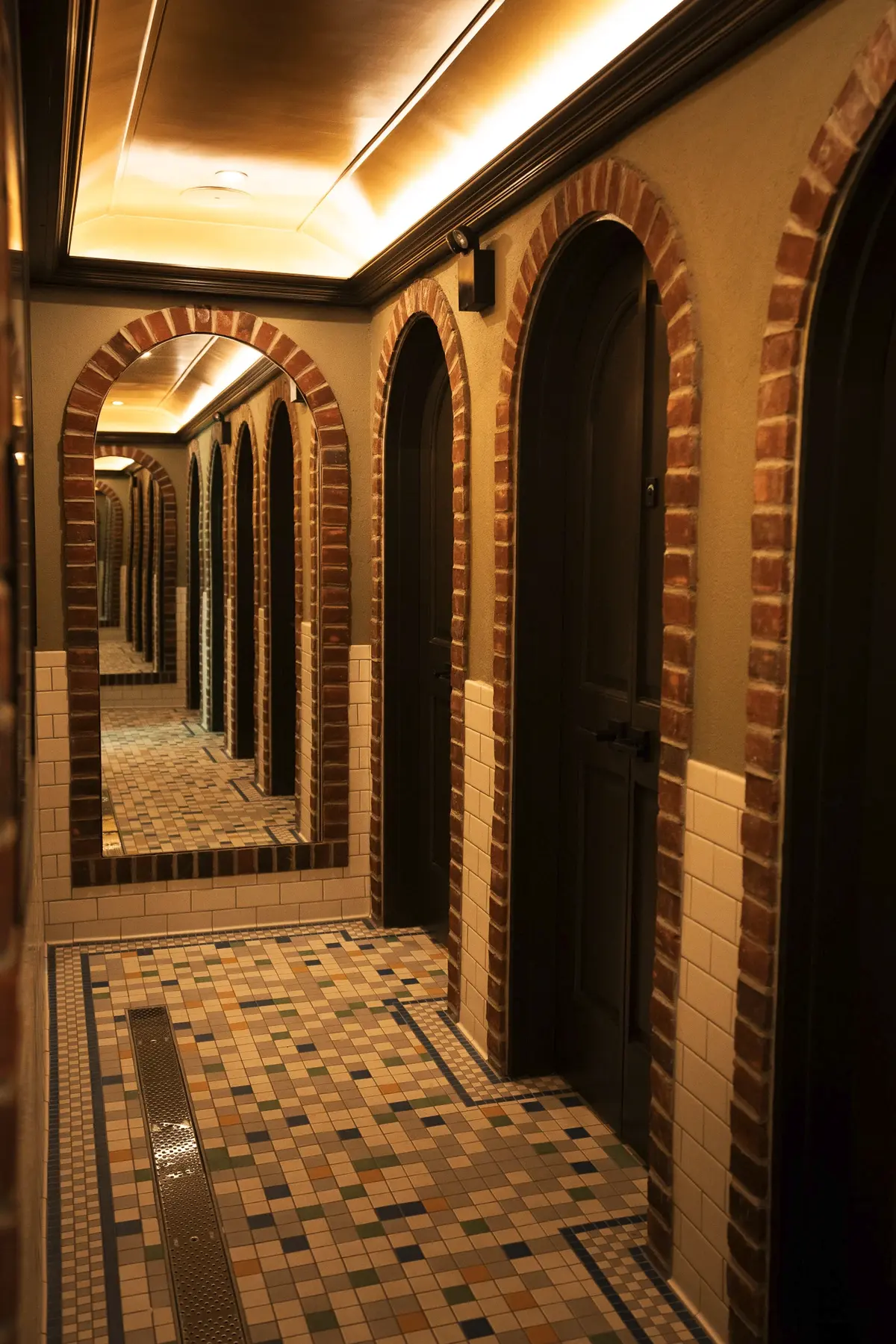 Brewery bathroom with mosaic floor tile, arched stall doors, brick-framed arch mirror, and coffered gold ceiling.